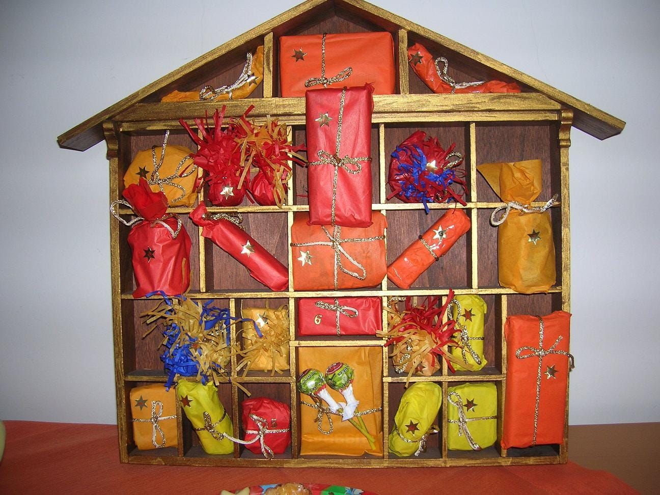 Holzregal in Hausform mit vielen Fächern voller kleiner Päckchen in rotem, orangem und gelbem Papier mit goldenen Sternen