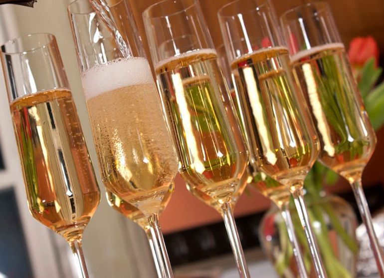 Mehrere Sektgläser in einer Reihe, eines wird gerade mit perlendem Sekt gefüllt, im Hintergrund rote Blumen