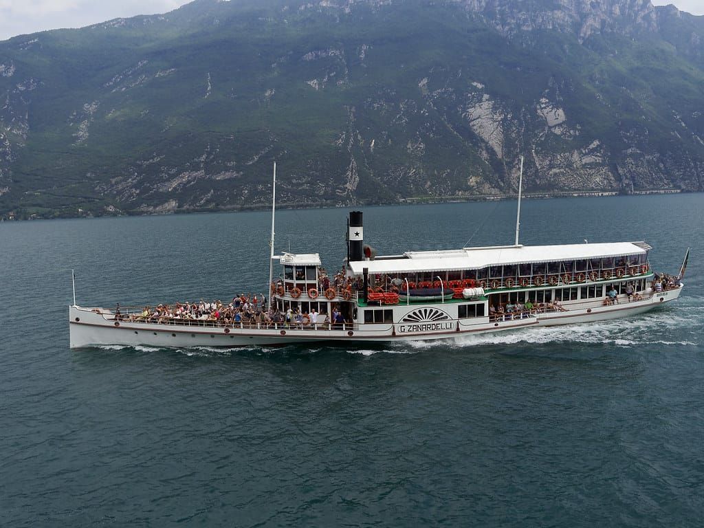 Weißer historischer Raddampfer mit Fahrgästen auf einem tiefblauen See vor steilen Bergen