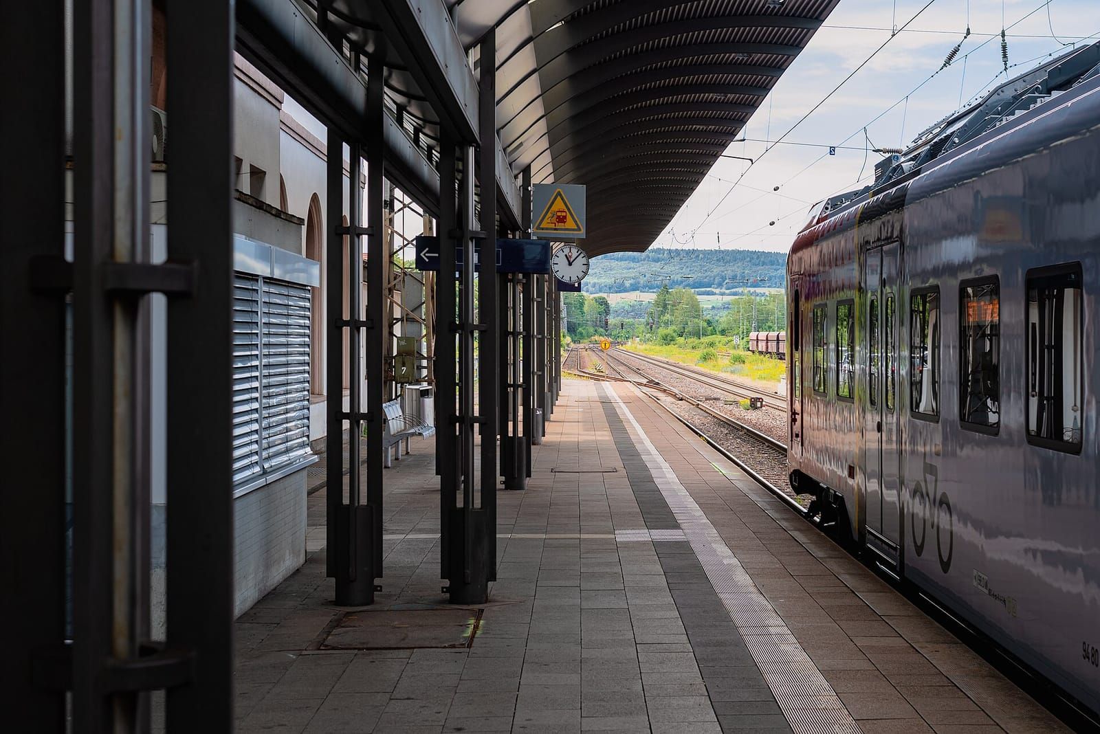 Überdachter Bahnsteig eines kleinen Bahnhofs mit Bahnhofsuhr, rechts ein silberner Regionalzug, im Hintergrund bewaldete Hügel