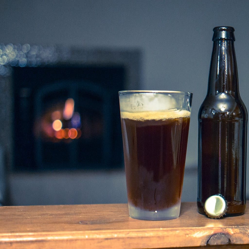 Glas mit dunklem, selbst gebrautem Bier und eine braune Flasche mit Kronkorken vor einem Kaminfeuer