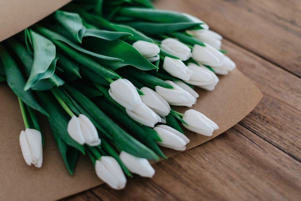 Strauß weißer Tulpen mit langen grünen Blättern, in braunes Packpapier eingeschlagen, auf einem Holztisch