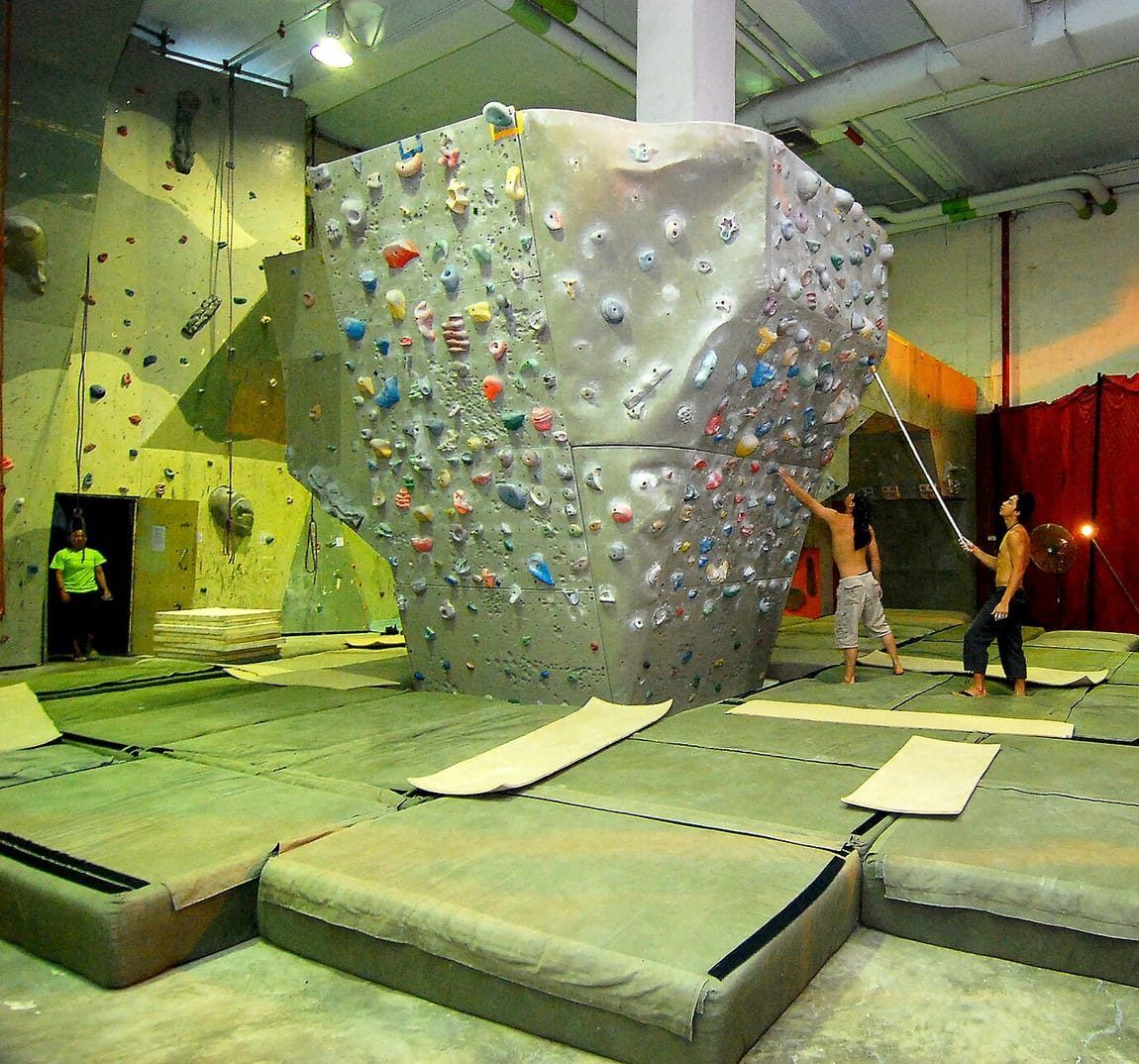 Boulderhalle mit großem grauem Kletterblock voller bunter Griffe und dicken grünen Weichbodenmatten