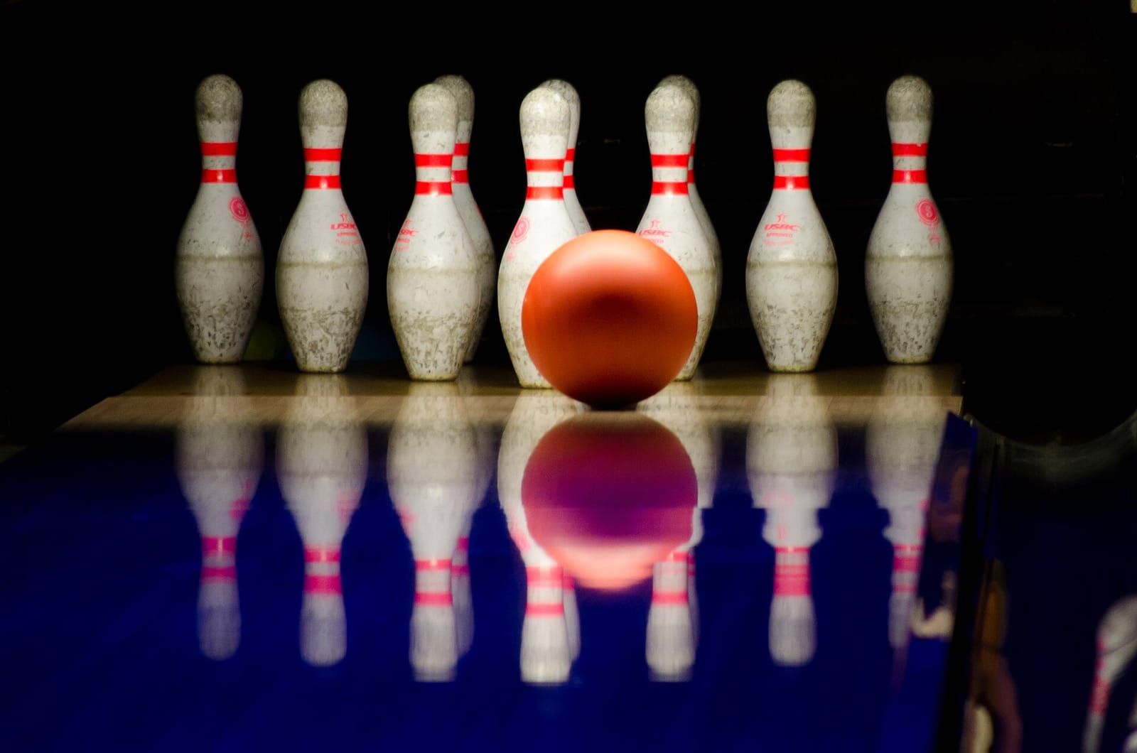 Orangefarbene Bowlingkugel rollt auf einer glänzenden Bahn auf zehn weiße Kegel mit roten Streifen zu