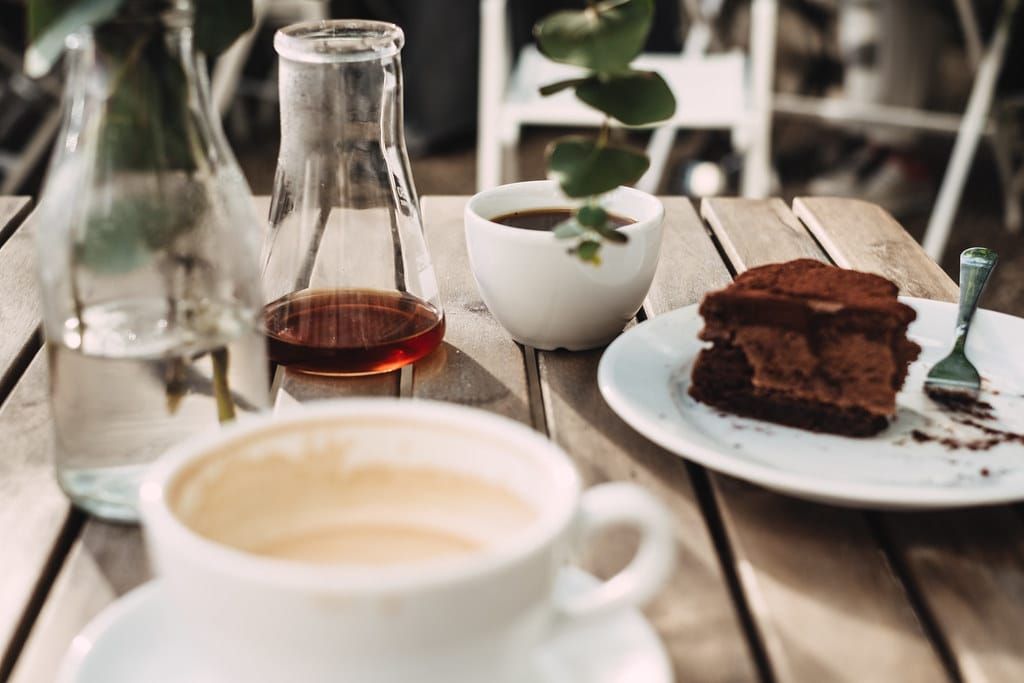 Holztisch in einem Café mit Cappuccinotasse, einem Stück Schokoladenkuchen auf einem Teller und einer Glaskaraffe