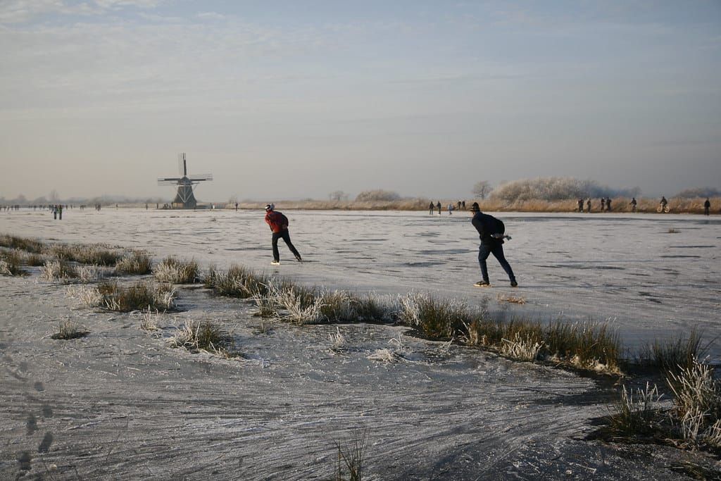 Zwei Eisläufer auf einem zugefrorenen See mit Raureif am Ufer, im Hintergrund eine Windmühle im Winterlicht