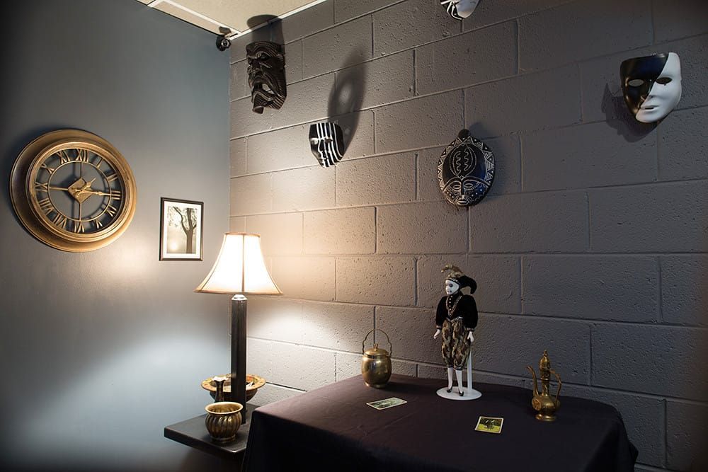 Gedämpft beleuchteter Raum mit Masken an einer Steinwand, einer alten Wanduhr, einer Lampe und einem Tisch mit Hinweiskarten
