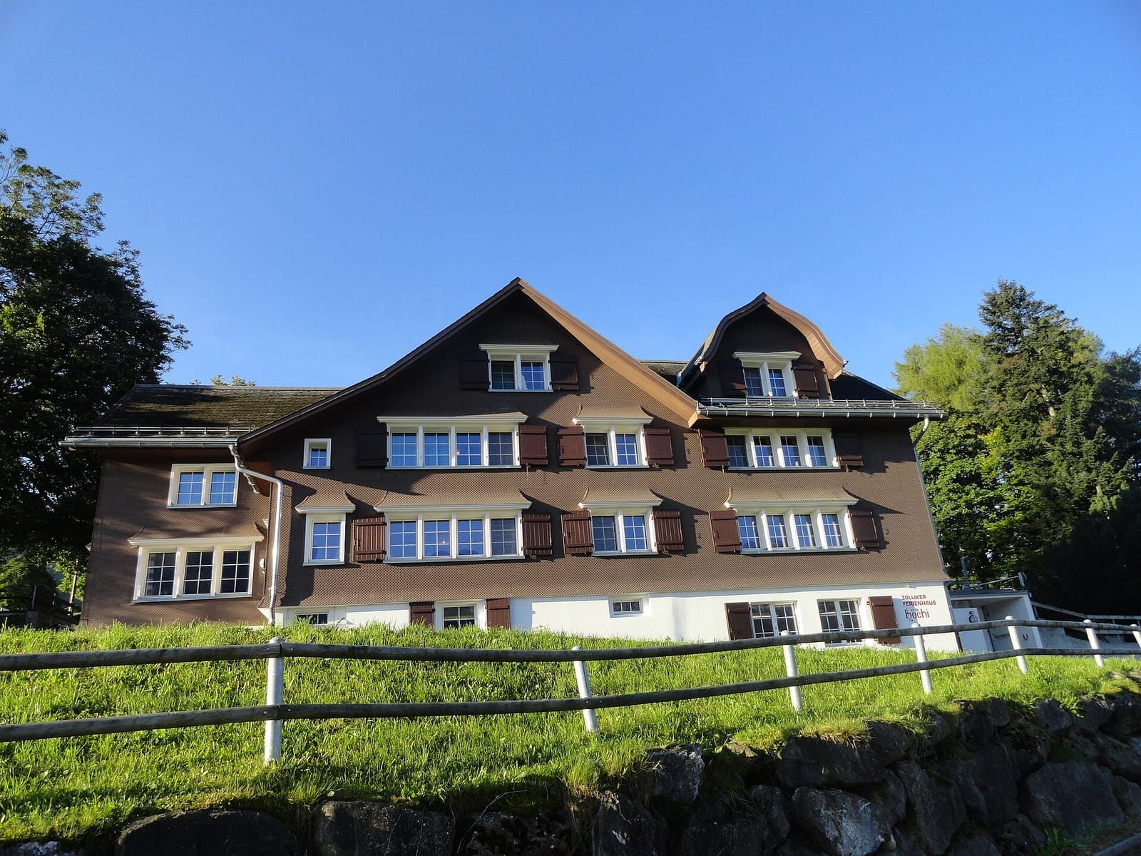 Großes Ferienhaus mit braun-weißer Holzfassade und vielen Fenstern auf einer grünen Wiese unter blauem Himmel