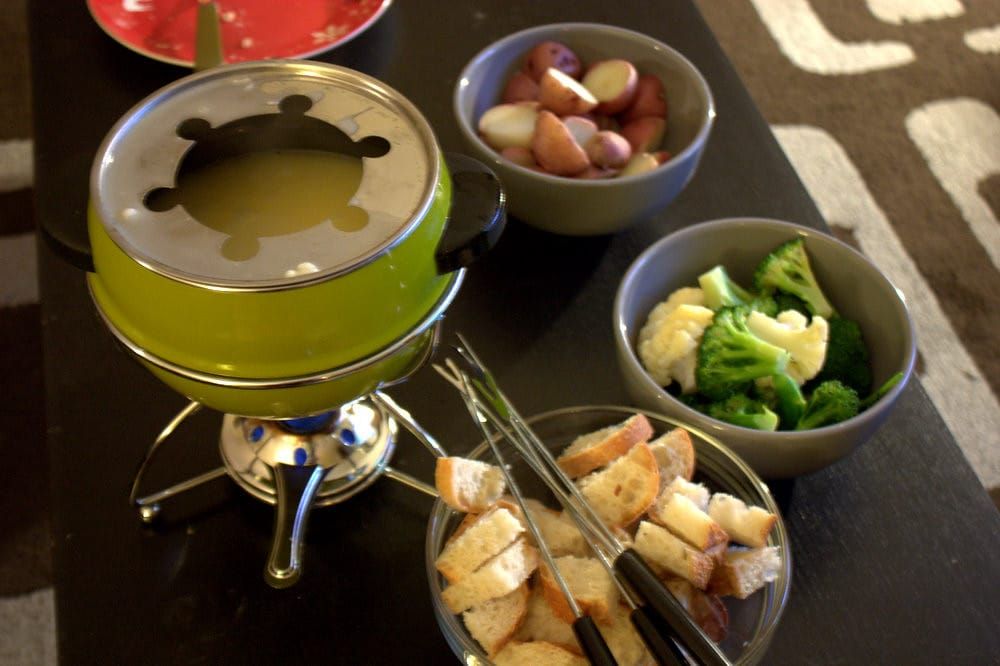 Grüner Fonduetopf auf einem Rechaud, daneben Schalen mit Brotwürfeln, Brokkoli, Blumenkohl und kleinen Kartoffeln