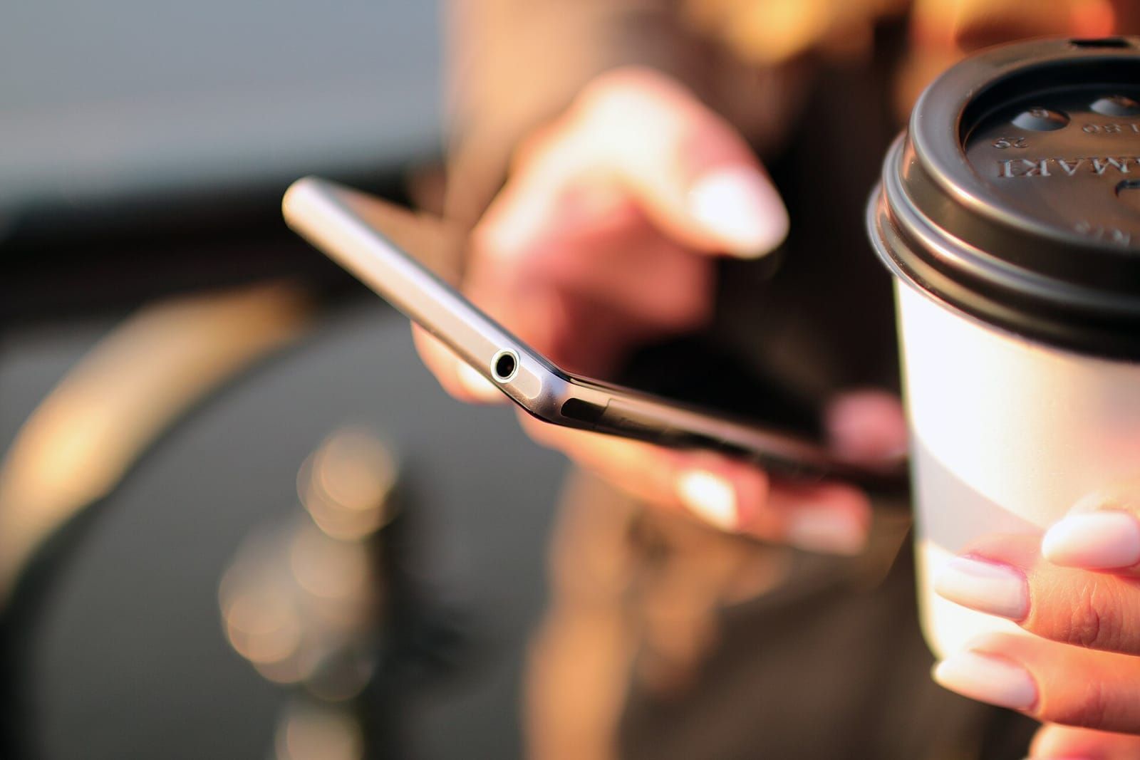 Hand tippt auf einem Smartphone, im Vordergrund ein Kaffeebecher zum Mitnehmen