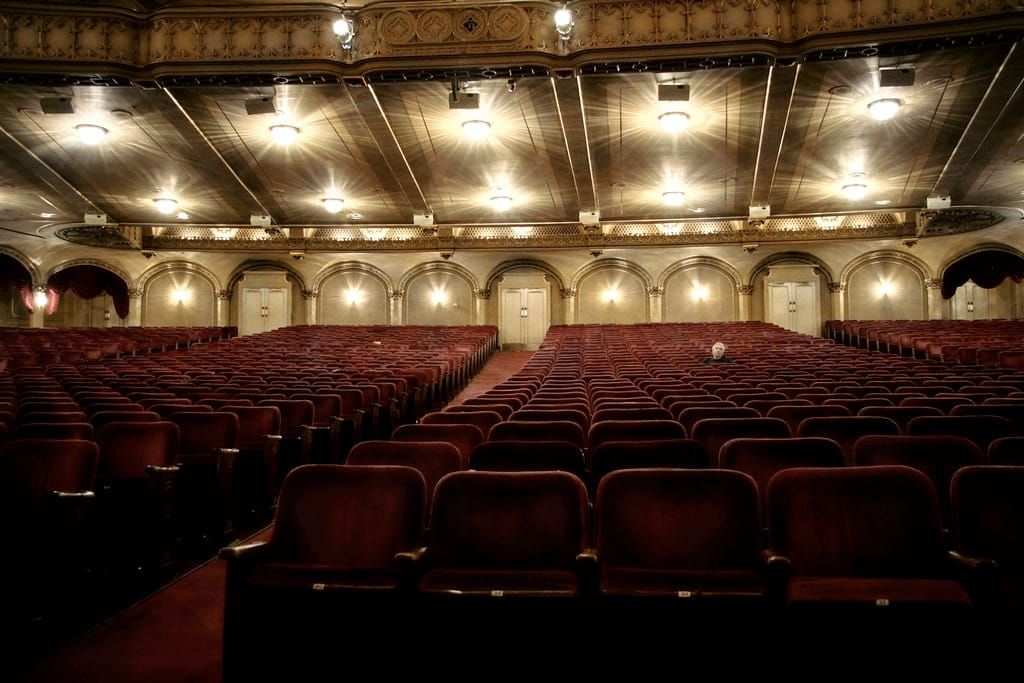 Großer, fast leerer Theatersaal mit roten Samtsitzreihen, goldverzierten Wänden und leuchtenden Deckenlampen