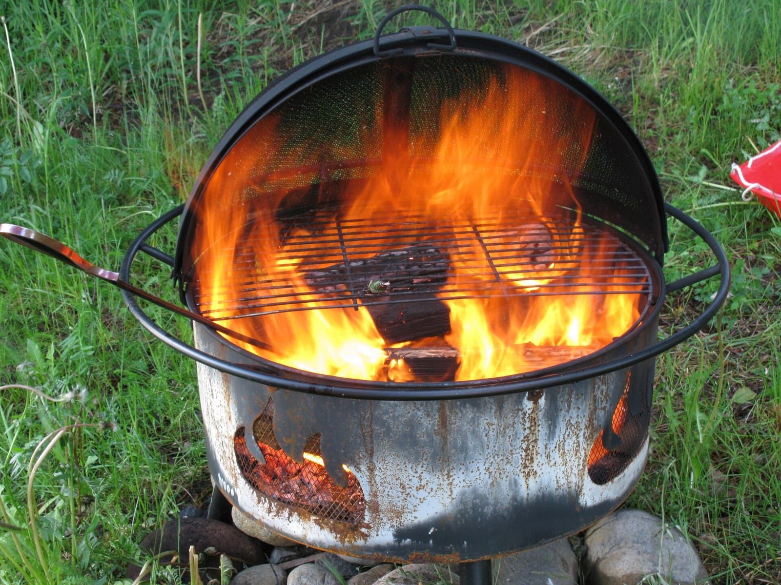 Runde Feuerschale aus Metall mit lodernden Flammen, Grillrost und Funkenschutz auf einer Wiese