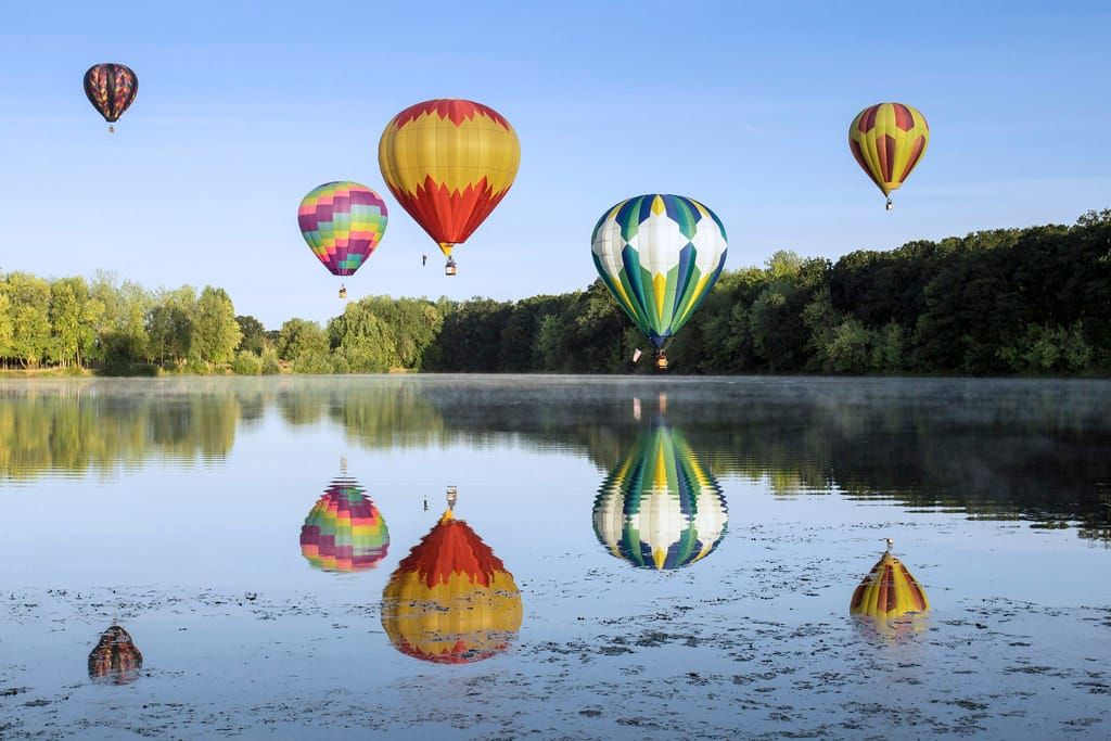Mehrere bunte Heißluftballons schweben über einem spiegelglatten See mit Waldrand an einem klaren Morgen