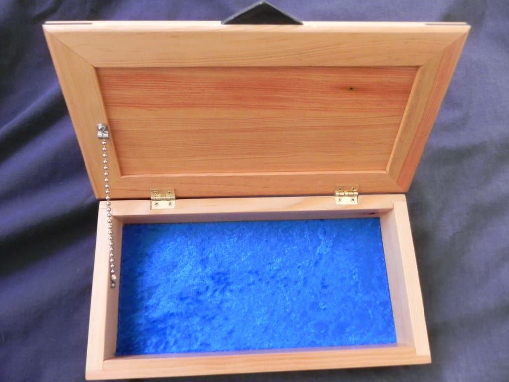 Aufgeklappte, leere Holzkiste mit blauem Samtfutter, Messingscharnieren und Kettchen auf dunklem Stoff