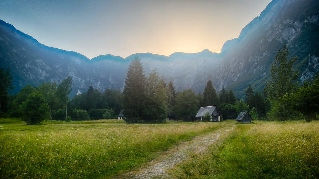 Grünes Hochtal mit einem Schotterweg zu zwei kleinen Holzhütten zwischen Nadelbäumen und steilen Felswänden
