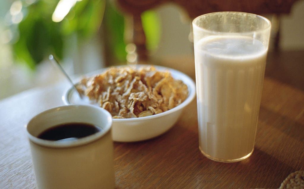 Tasse schwarzer Kaffee, Schale mit Cornflakes und ein Glas Milch auf einem Holztisch im Morgenlicht