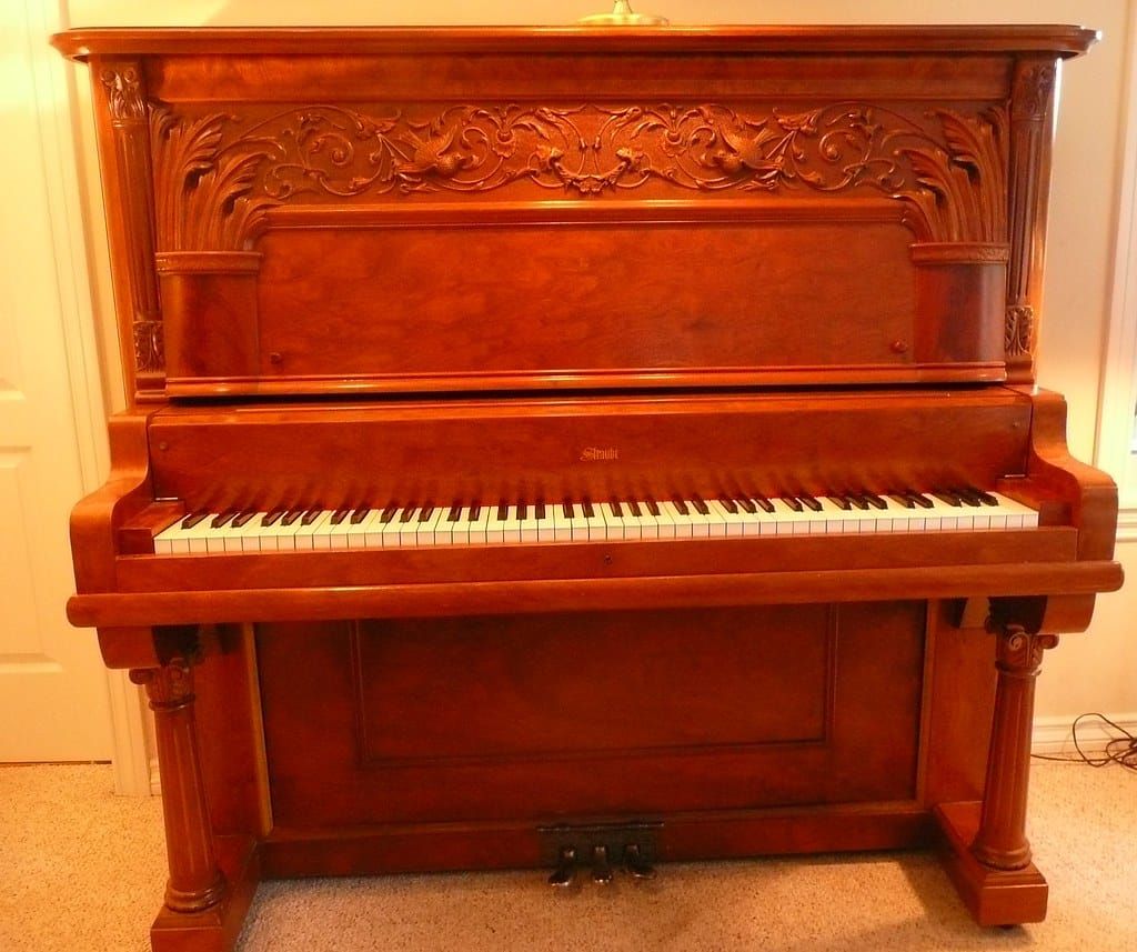 Altes Klavier aus rötlichem Holz mit geschnitzten Ornamenten in einem hellen Wohnzimmer