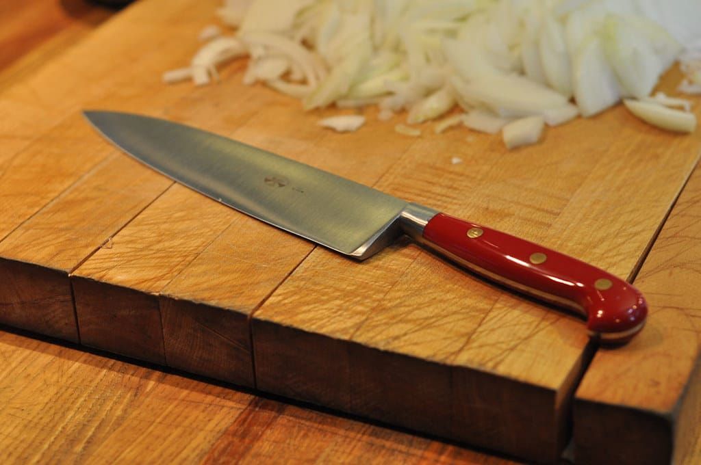 Großes Kochmesser mit rotem Griff und Messingnieten auf einem Holzbrett neben geschnittenen Zwiebeln
