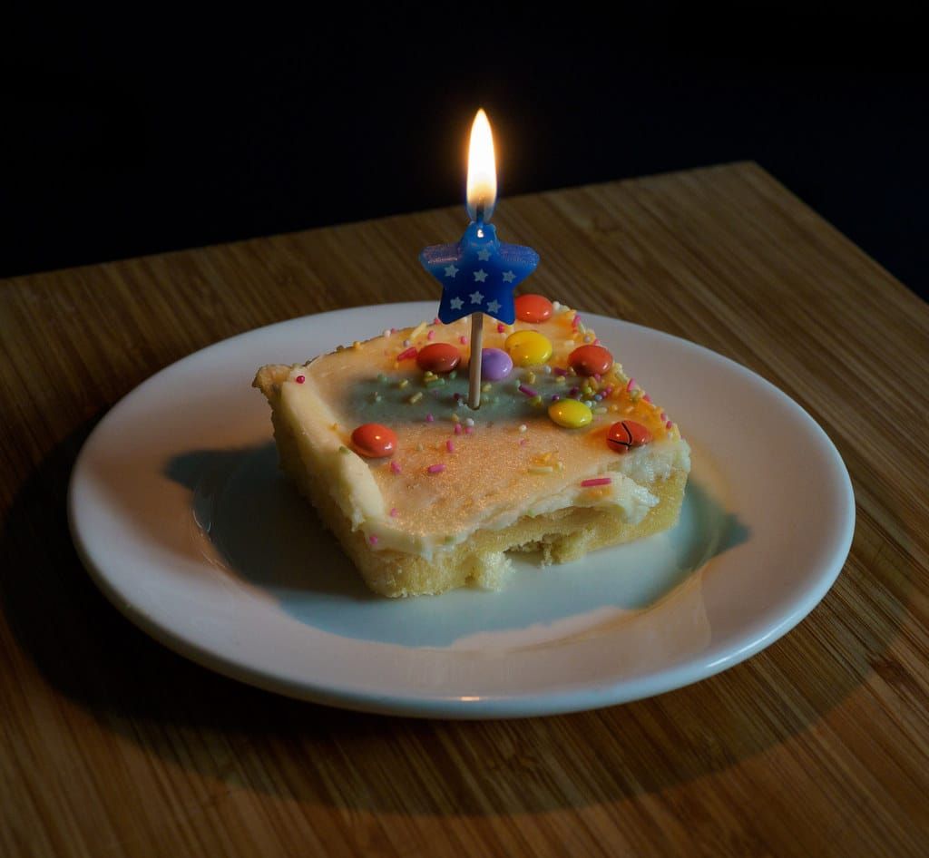 Stück Rührkuchen mit Zuckerguss, bunten Schokolinsen und Streuseln auf einem Teller, darin eine brennende blaue Sternkerze