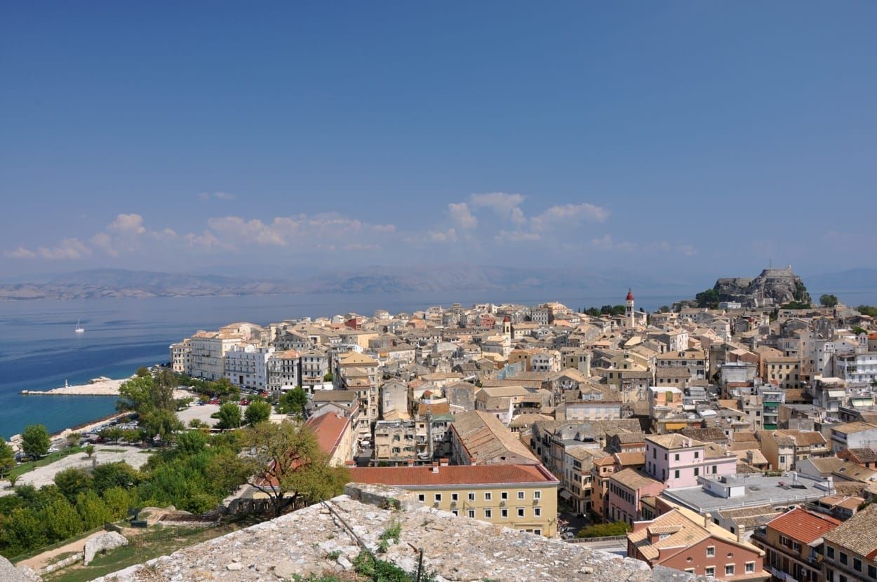 Blick über die Altstadt von Korfu mit hellen Häusern, Kirchturm und Meer unter blauem Himmel