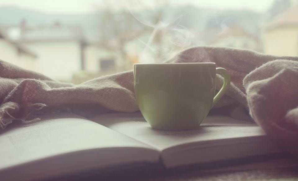 Grüne Tasse mit dampfendem Getränk auf einem aufgeschlagenen Buch, daneben eine graue Wolldecke am Fenster