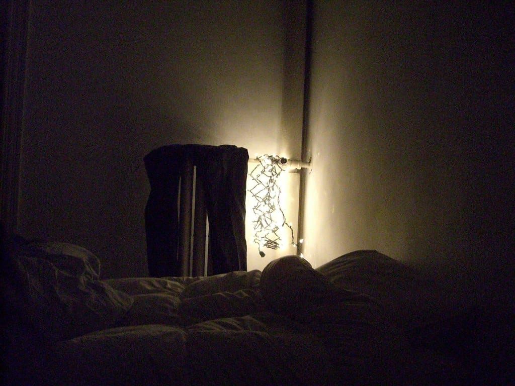 Warmweiße Lichterkette hängt in einer dunklen Zimmerecke an einem Heizungsrohr neben einem Bett