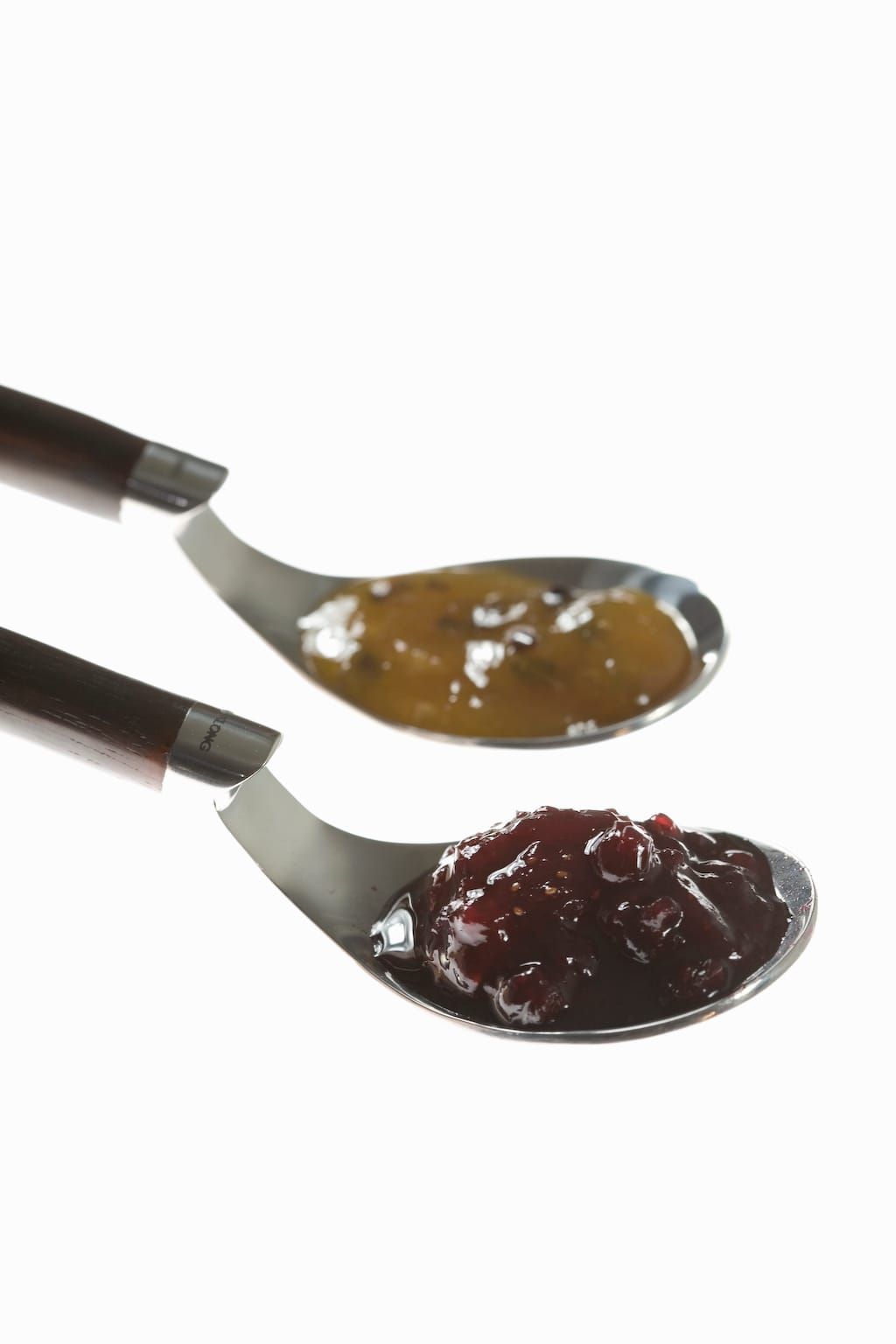 Zwei Metalllöffel mit dunkelroter Beerenmarmelade und orangefarbener Konfitüre vor weißem Hintergrund