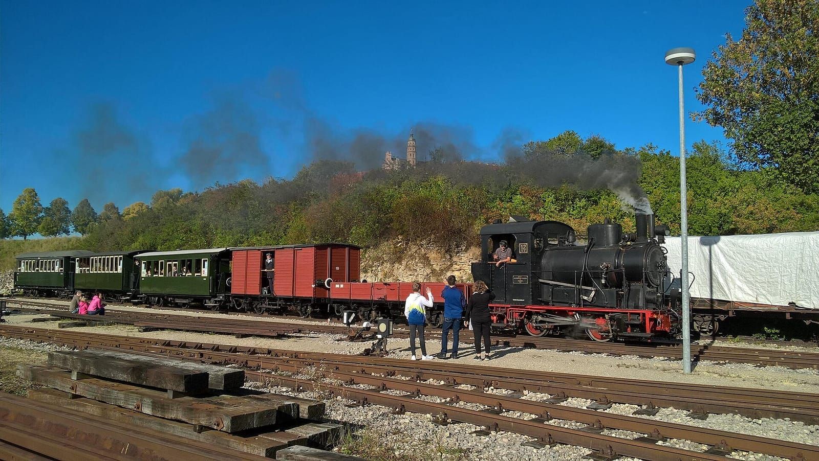 Historische Dampflok mit roten und grünen Waggons auf einem Bahnhof, dunkler Rauch vor blauem Himmel