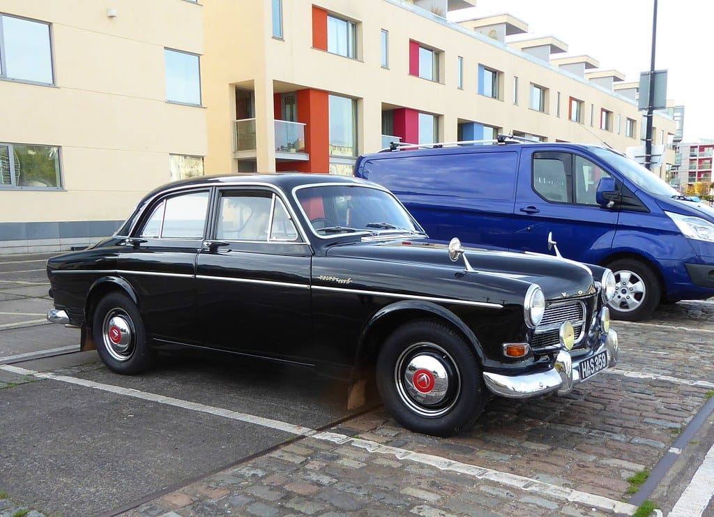 Schwarzer Oldtimer aus den Sechzigerjahren mit Chromstoßstange und hellen Radzierringen auf einem Pflasterparkplatz