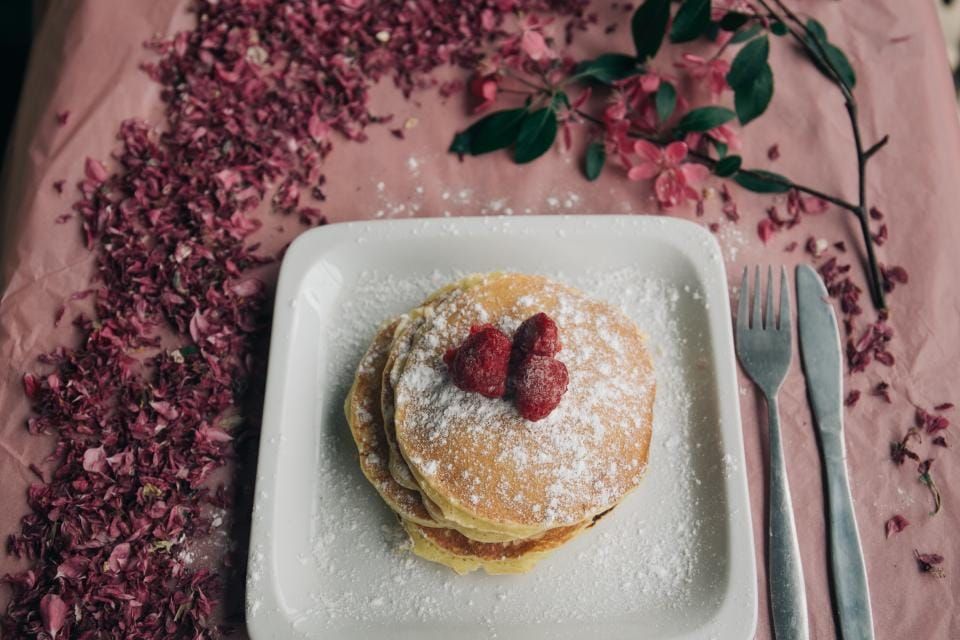 Stapel Pfannkuchen mit Puderzucker und Himbeeren auf einem weißen Teller, umgeben von rosa Blütenblättern