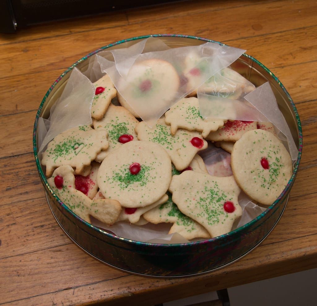 Runde Blechdose mit selbstgebackenen Butterplätzchen, verziert mit grünem Zucker und roten Bonbons, auf einem Holztisch