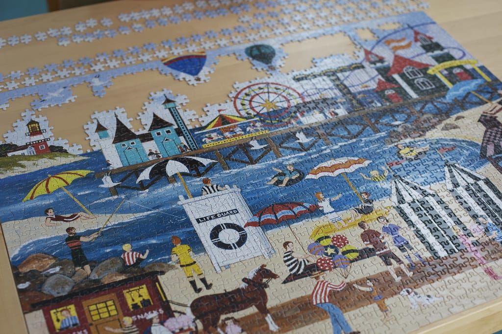 Halb fertiges Puzzle mit bunter Strandszene, Riesenrad und Leuchtturm auf einem hellen Holztisch