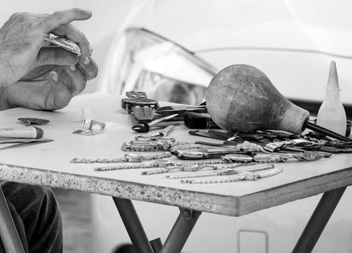 Schwarzweißfoto: Hände eines Uhrmachers über einem Klapptisch mit Armbanduhren, Uhrenarmbändern, Pinsel und Blasebalg
