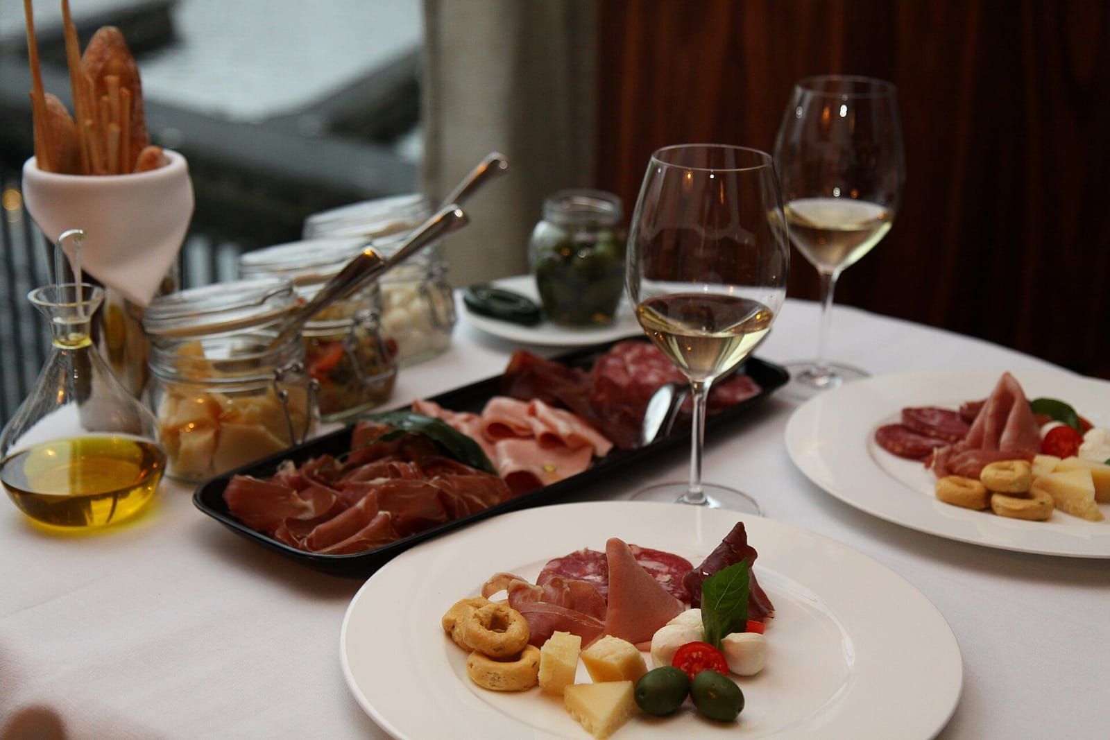Gedeckter Restauranttisch mit Antipasti-Tellern, Schinken, Käse, Oliven und zwei Gläsern Weißwein