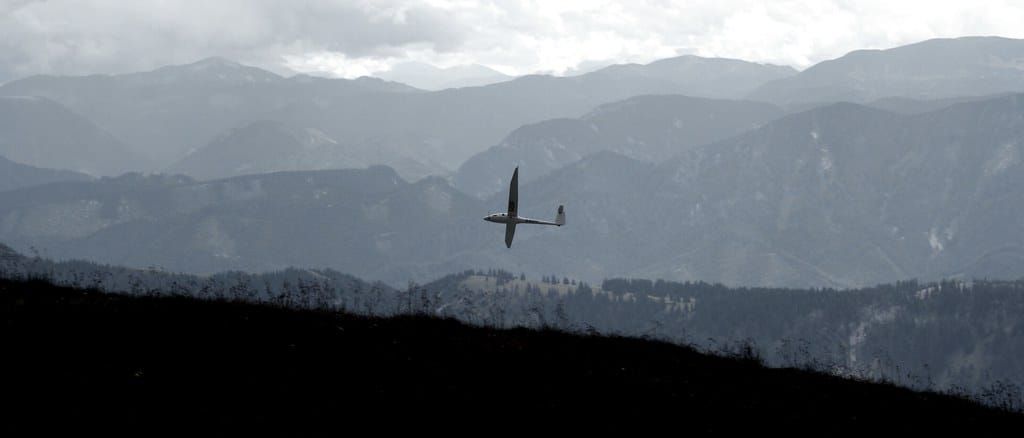Segelflugzeug mit langen schmalen Tragflächen fliegt vor einer dunstigen Bergkette über bewaldeten Hängen