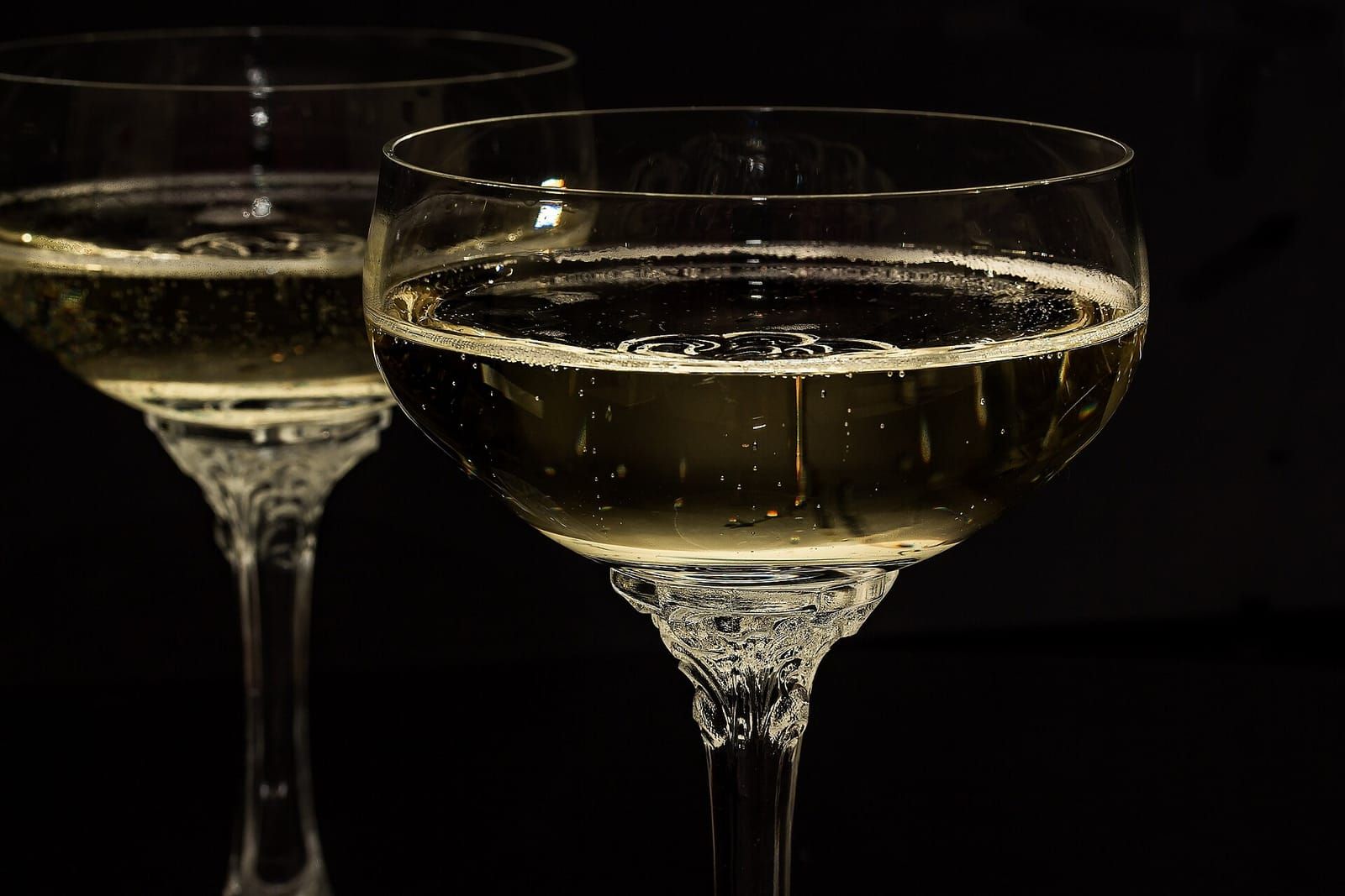 Zwei Sektschalen aus Kristallglas mit verziertem Stiel, gefüllt mit Sekt, vor schwarzem Hintergrund