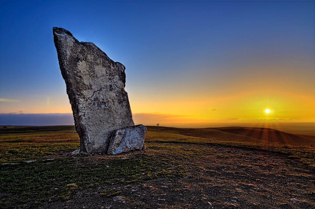 Hoher, schmaler Felsen auf einer weiten Ebene, im Hintergrund geht die Sonne am Horizont unter einem klaren blauen Himmel auf