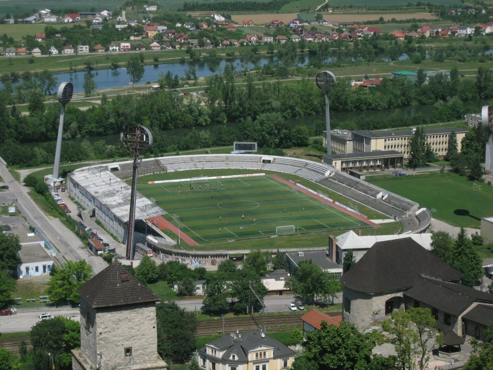 Luftaufnahme eines Fußballstadions mit Flutlichtmasten und Tribünen am Rand einer Kleinstadt