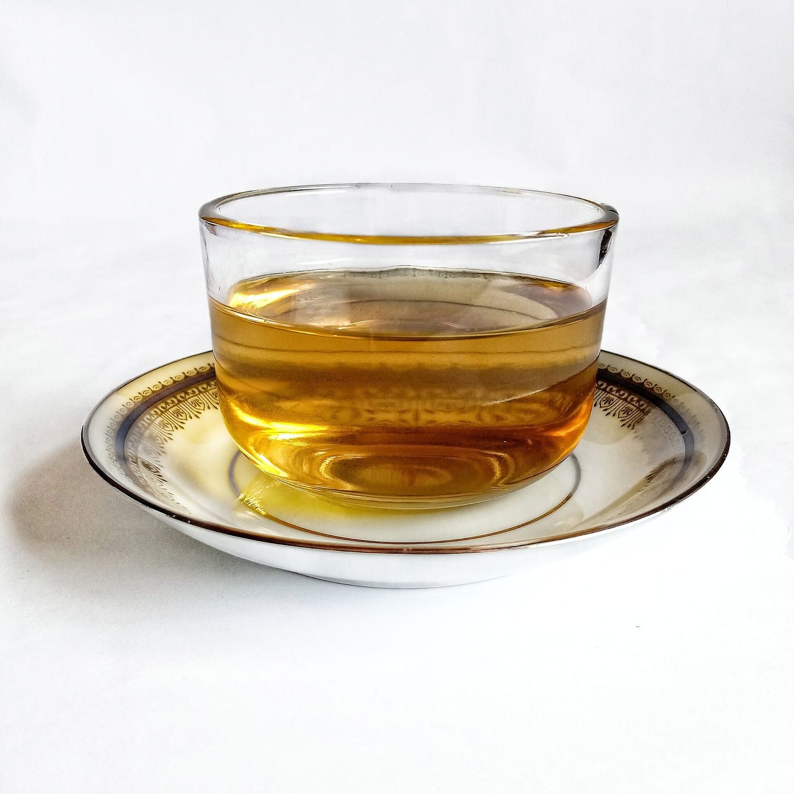 Gläserne Teeschale mit goldgelbem Tee auf einer weißen Untertasse mit Goldrand vor hellem Hintergrund