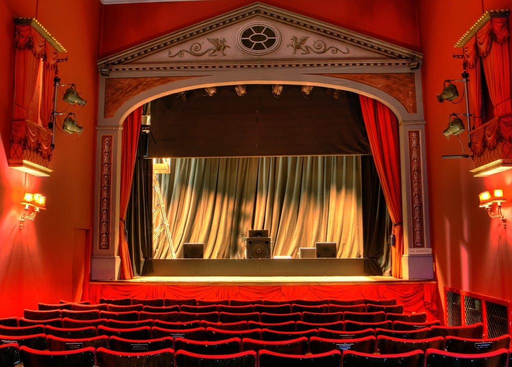 Kleine Theaterbühne mit roten Vorhängen, Scheinwerfern und leeren roten Sitzreihen davor