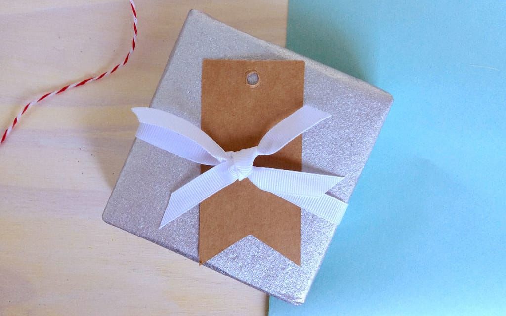 Kleines, silbern verpacktes Geschenk mit weißer Schleife und braunem Papieranhänger auf einem Holztisch