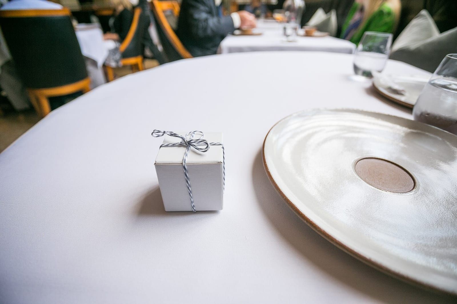 Kleine weiße Geschenkschachtel mit grau-weißer Kordel auf einem gedeckten Tisch neben einem Teller