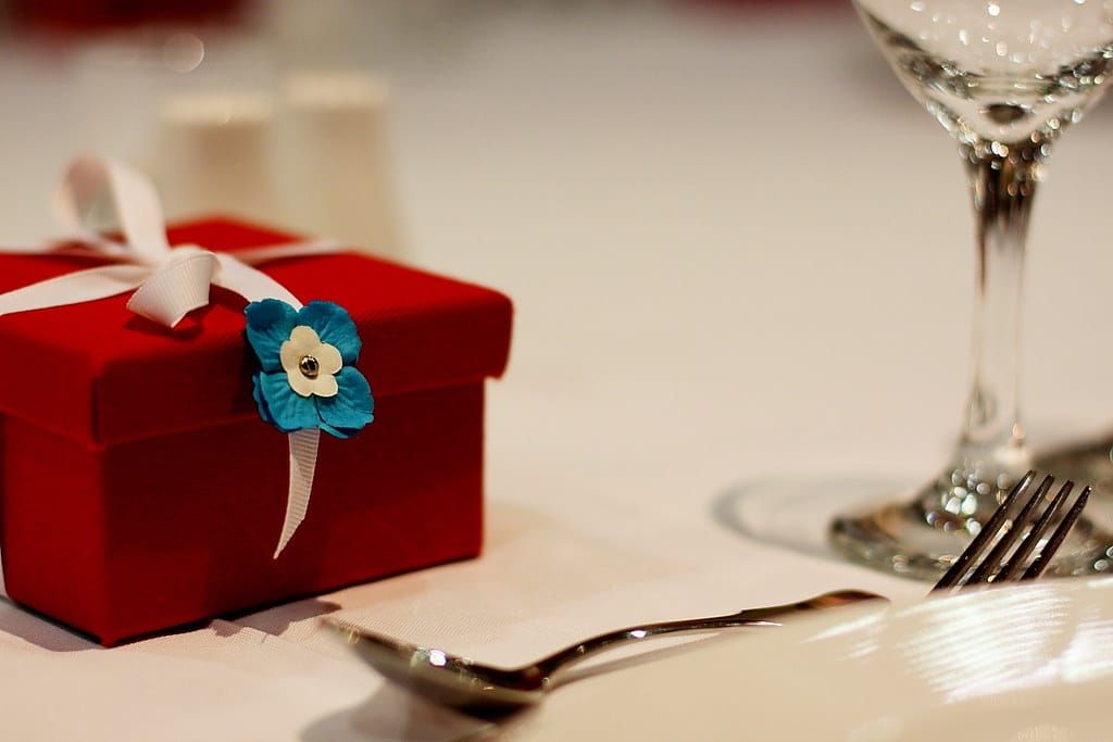 Rote Geschenkschachtel mit weißer Schleife und blauer Papierblüte auf einem gedeckten Tisch neben Weinglas und Besteck
