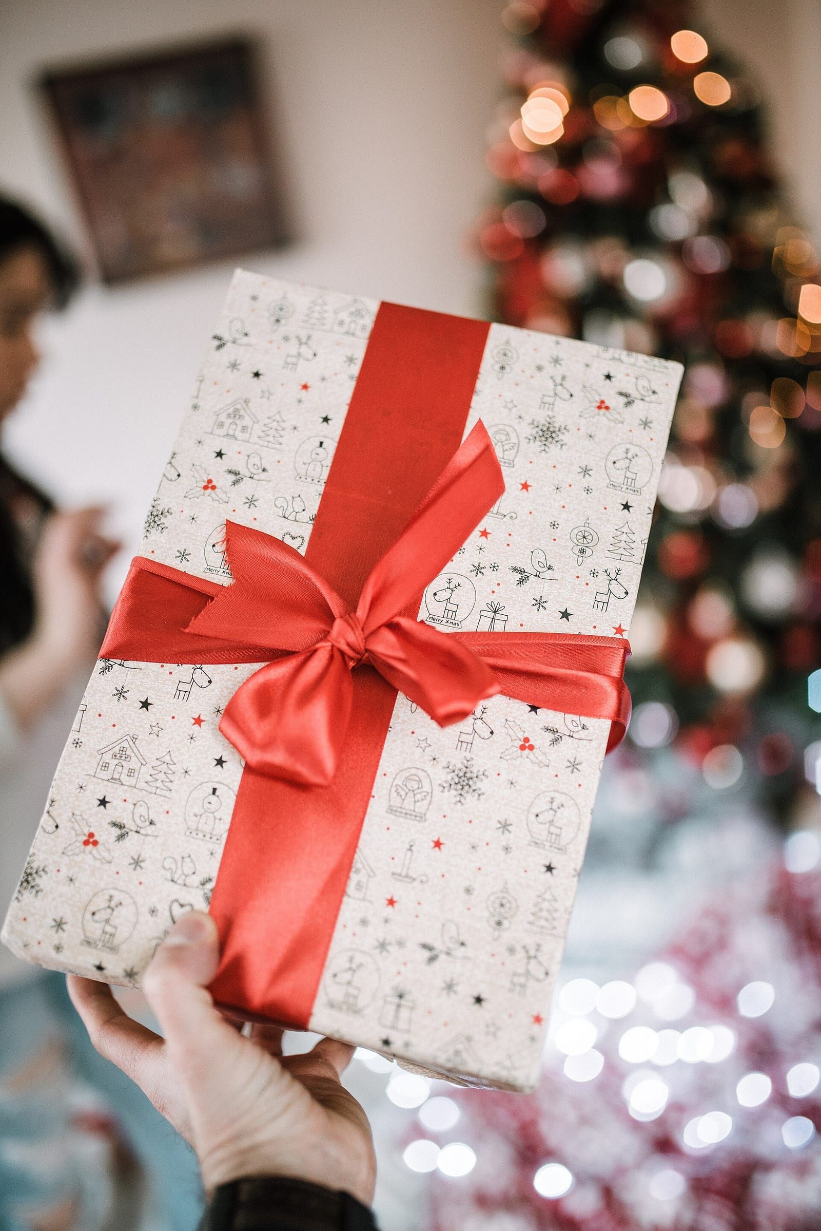 Eine Hand hält ein Geschenk mit roter Schleife vor einem unscharf leuchtenden Weihnachtsbaum