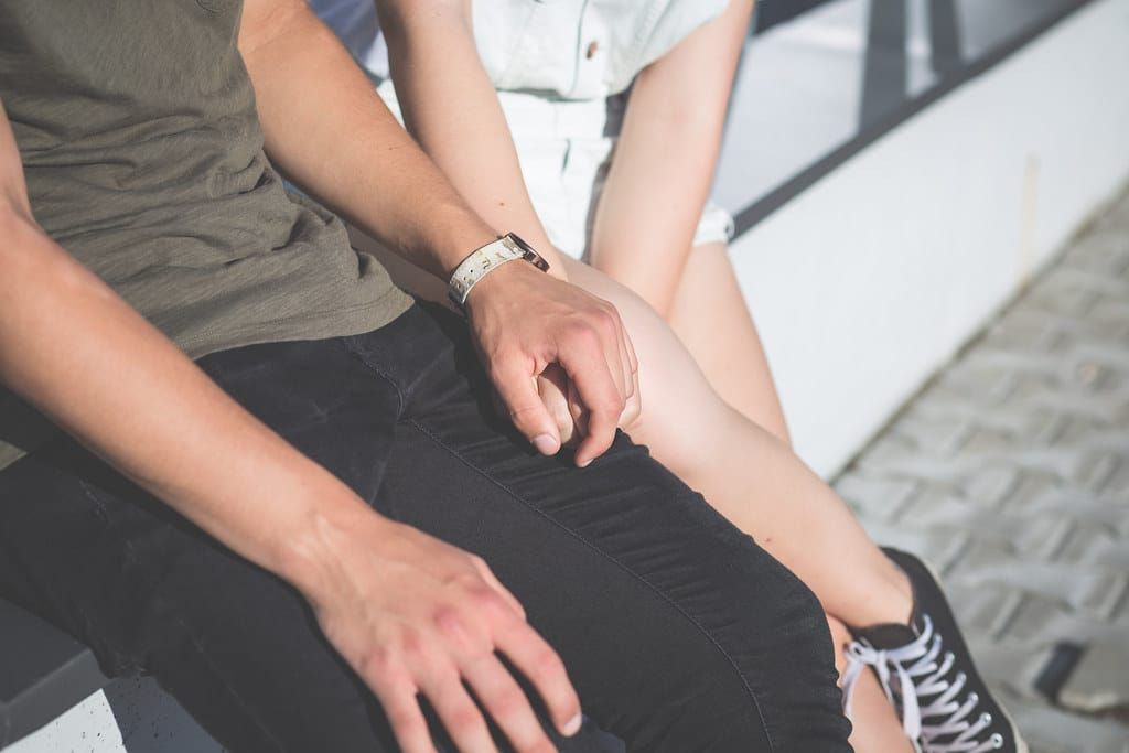 Ein Paar sitzt nebeneinander auf einer Bank, nur Arme und Beine sind zu sehen, er hält ihre Hand auf seinem Knie