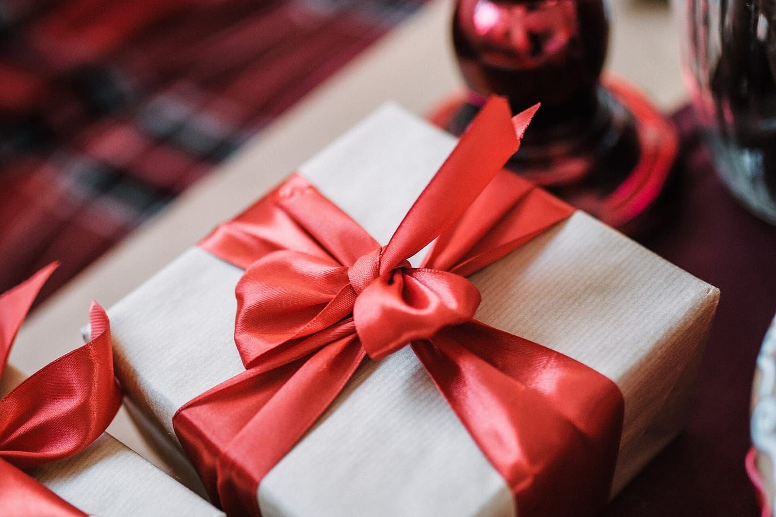 In hellbraunes Papier eingepacktes Geschenk mit roter Satinschleife auf einem Tisch, daneben ein rotes Glas und ein zweites Päckchen