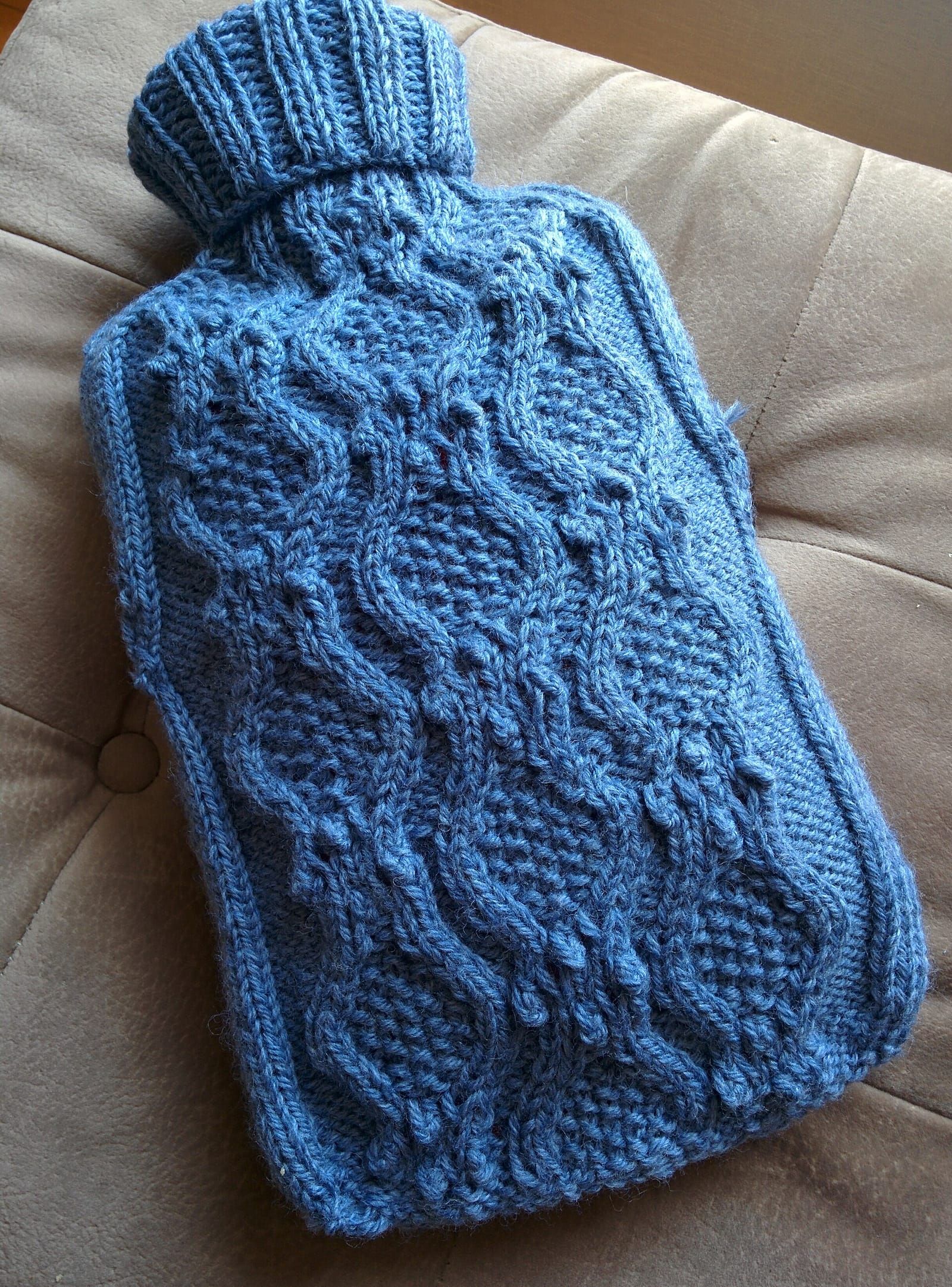 Wärmflasche in einem blauen Strickbezug mit Zopfmuster, liegend auf einem beigefarbenen Sofakissen