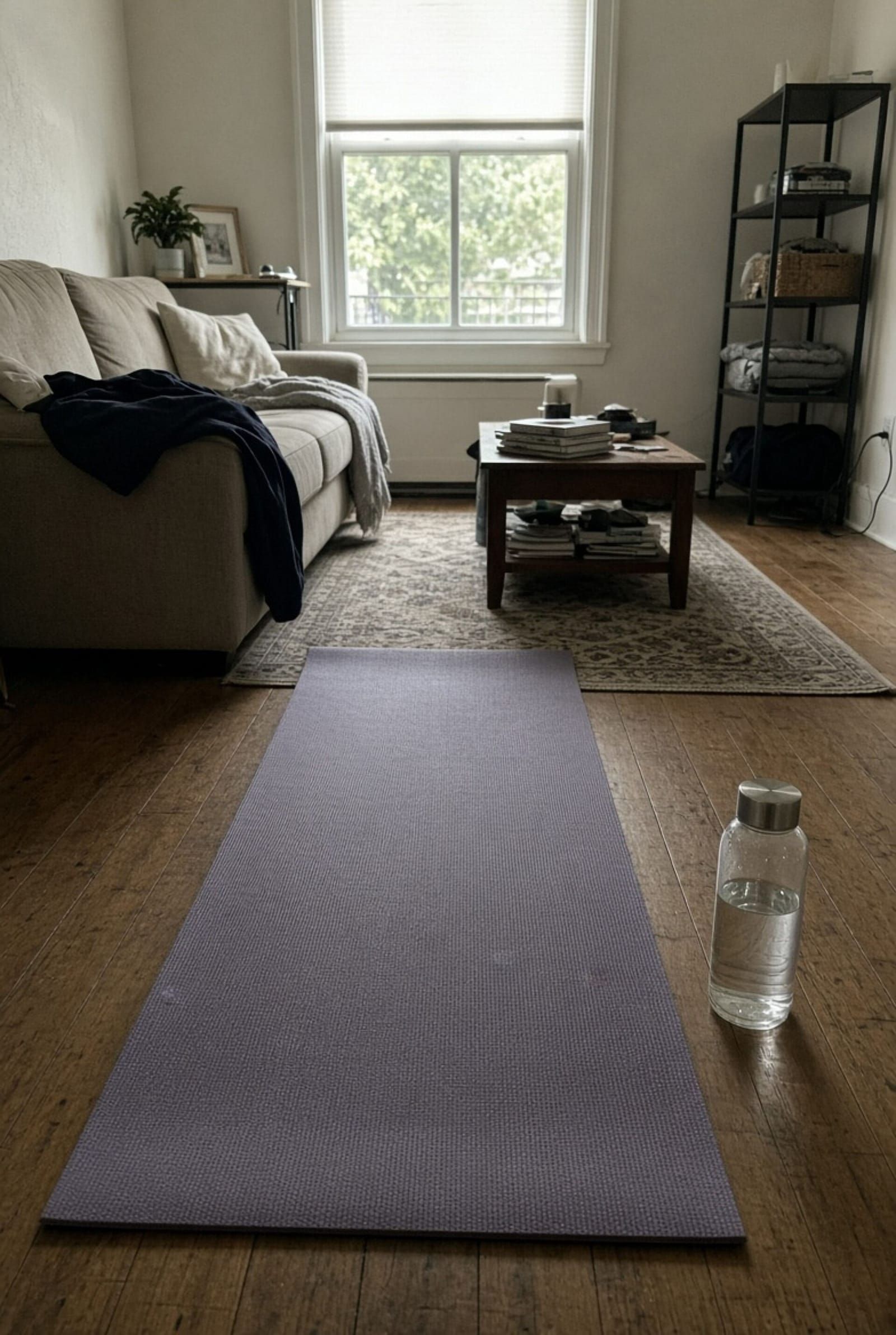 Ausgerollte violette Yogamatte auf einem Holzboden, daneben eine Wasserflasche, im Hintergrund Sofa und Fenster
