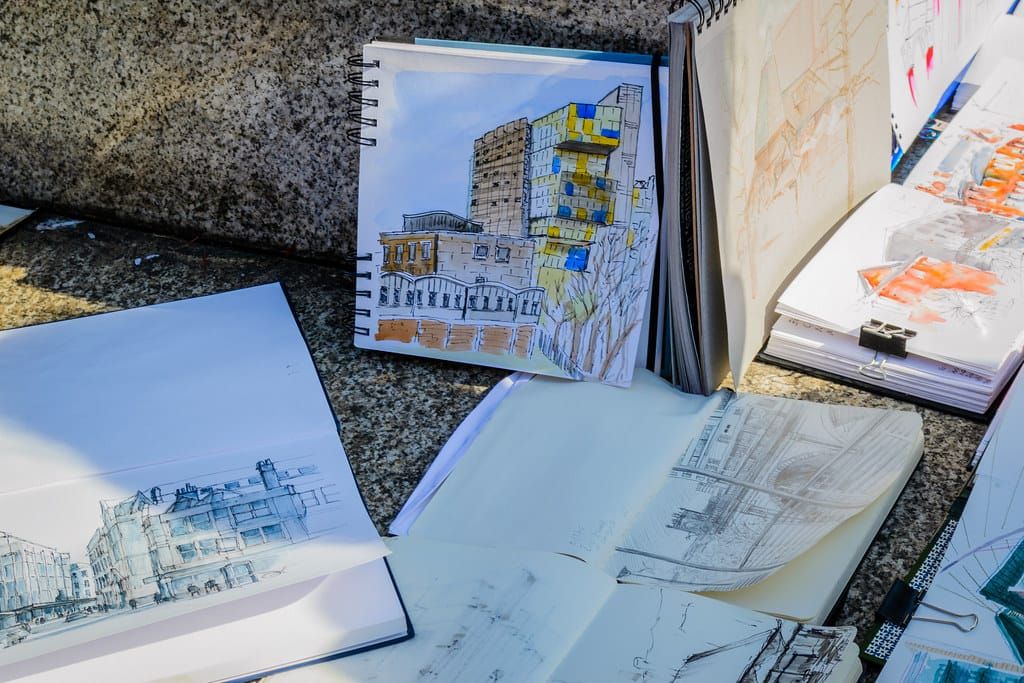 Mehrere aufgeschlagene Skizzenbücher mit farbigen Zeichnungen von Stadthäusern und Gebäuden auf einer Steinbank