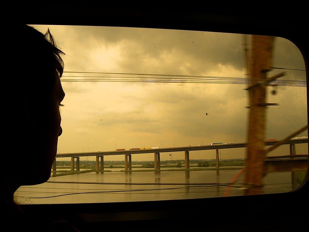 Silhouette eines Kopfes vor einem Zugfenster, draußen eine lange Brücke über Wasser unter goldgelbem Himmel